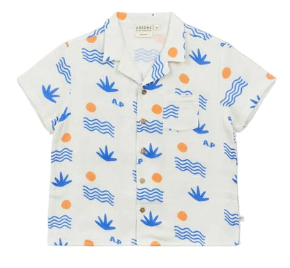 The Arsene & Les Pipelettes Boys' Magnum Shirt is a short-sleeve white double voile shirt featuring blue wave and plant motifs, orange sun accents, a chest pocket, and brown buttons, showcased against a plain background.