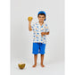 A young boy in blue shorts and a blue cap stands barefoot near a coconut on a plain white background, holding a yellow toy and wearing the Arsene & Les Pipelettes Boys' Magnum Shirt.