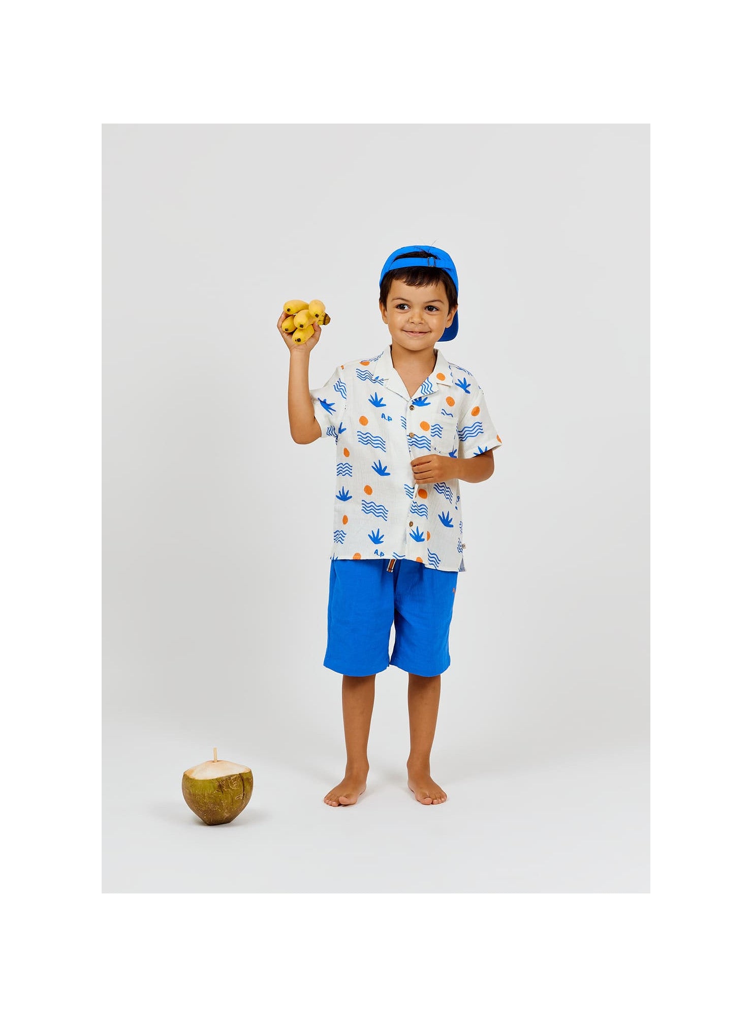 A young boy in blue shorts and a blue cap stands barefoot near a coconut on a plain white background, holding a yellow toy and wearing the Arsene & Les Pipelettes Boys' Magnum Shirt.