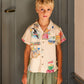 A young boy with blond hair is wearing the Arsene & Les Pipelettes Boys' Magnum Shirt, a relaxed fit, short sleeve cotton shirt with colorful prints, paired with green pants, as he stands indoors in front of a closed door.