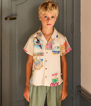 A young boy with blond hair is wearing the Arsene & Les Pipelettes Boys' Magnum Shirt, a relaxed fit, short sleeve cotton shirt with colorful prints, paired with green pants, as he stands indoors in front of a closed door.