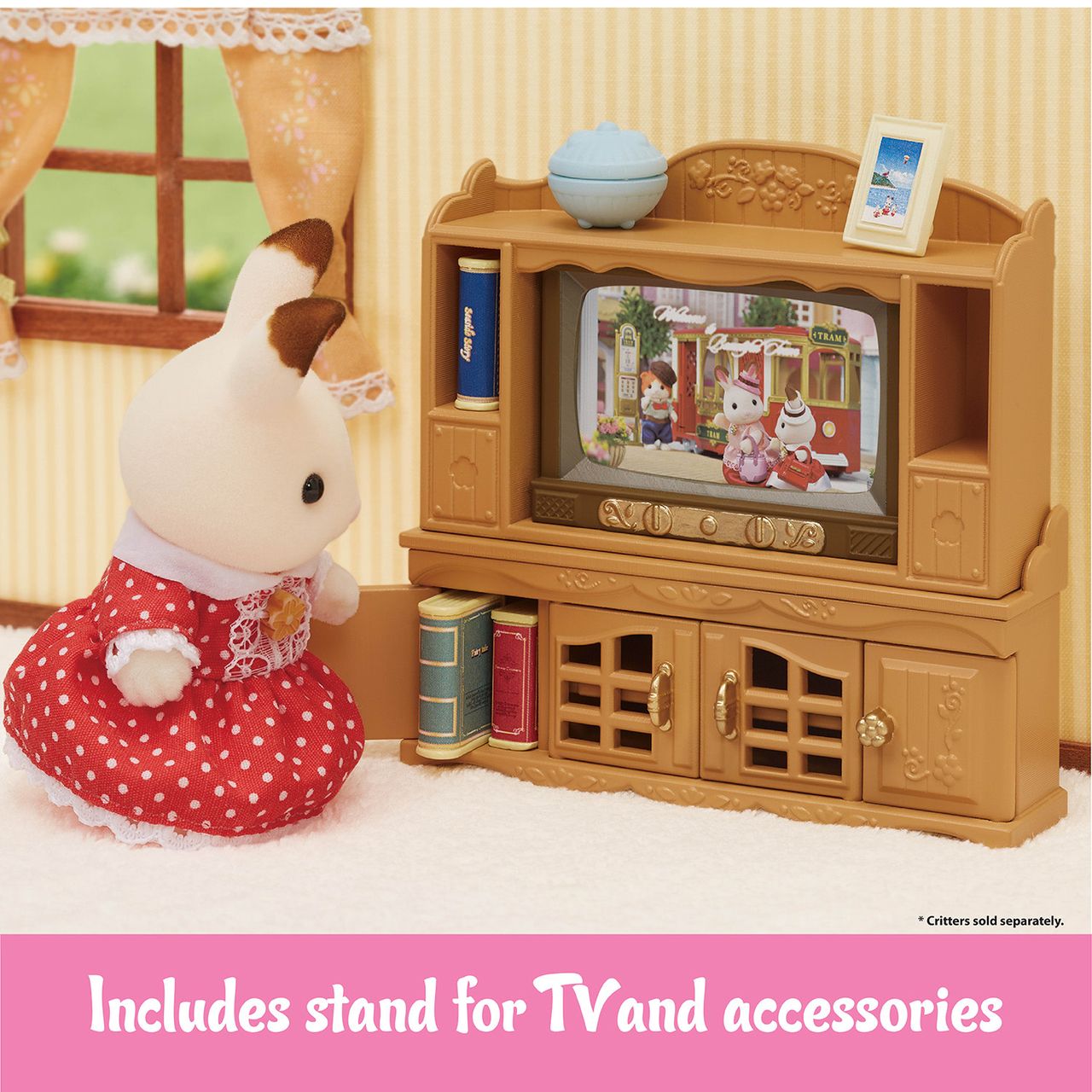 A Calico Critters bunny dressed in red sits in front of the Comfy Living Room Set, which features a TV stand with a small television, books, a globe, and a framed picture—ideal for your Calico Critters living room. Text at the bottom reads: “Includes stand for TV and accessories.”.