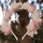The Noralee Pom Pom Crown + Wand, adorned with pink and cream tulle flowers, hangs on a branch—perfect for dress-up play—with blurred trees in the background.