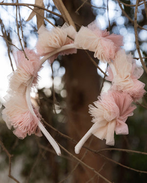The Noralee Pom Pom Crown + Wand, adorned with pink and cream tulle flowers, hangs on a branch—perfect for dress-up play—with blurred trees in the background.