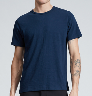 A person wearing the Rag & Bone Classic Flame Tee, a plain navy blue short-sleeve T-shirt crafted from durable Peruvian cotton, along with black pants and displaying tattoos on both forearms, is standing against a neutral background.