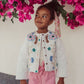 A girl wearing Louise Misha's Girls' Amandine Pant and a white floral embroidered jacket stands under pink flowers, gazing sideways.