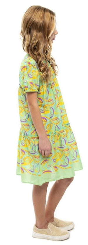 A young girl with long blonde hair stands in profile, wearing a Briton Court Girls' Meadows Kit Dress adorned with a colorful abstract pattern and beige shoes, made of lightweight cotton.