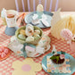 A pastel Easter table setting includes eggs with Meri Meri's Easter Icons Egg Decorating Tattoo Set, a pink tablecloth, bunny decorations, and a cupcake on a flower-shaped plate.