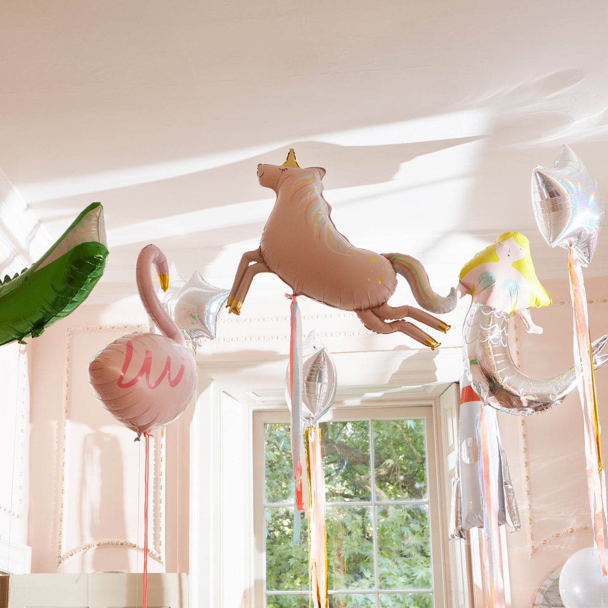 A set of Meri Meri Foil Balloons, featuring designs like a unicorn, flamingo, mermaid, star, and banana, floats near a window—ideal party decorations to brighten up any celebration.