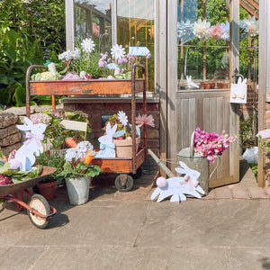 Outside a greenhouse, Meri Meri's Egg Hunt Kit adds charm with dapper bunny signs. Various plants and flowers are beautifully arranged on carts, baskets, and pots on a stone patio, creating an ideal setting for an Easter egg hunt.