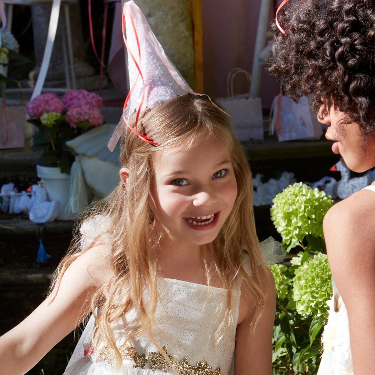 A young girl wearing a Meri Meri Party Hat smiles at another person at an outdoor event, surrounded by lush flowers and playful dinosaur decorations in the background.