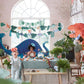 Three children wearing paper hats are at a Dinosaur Kingdom-themed party with colorful decorations, large dinosaur figures, and a table set with snacks and drinks. A Meri Meri Dinosaur Kingdom Large Garland by Meri Meri adds to the festive atmosphere as one child stretches their arms excitedly.