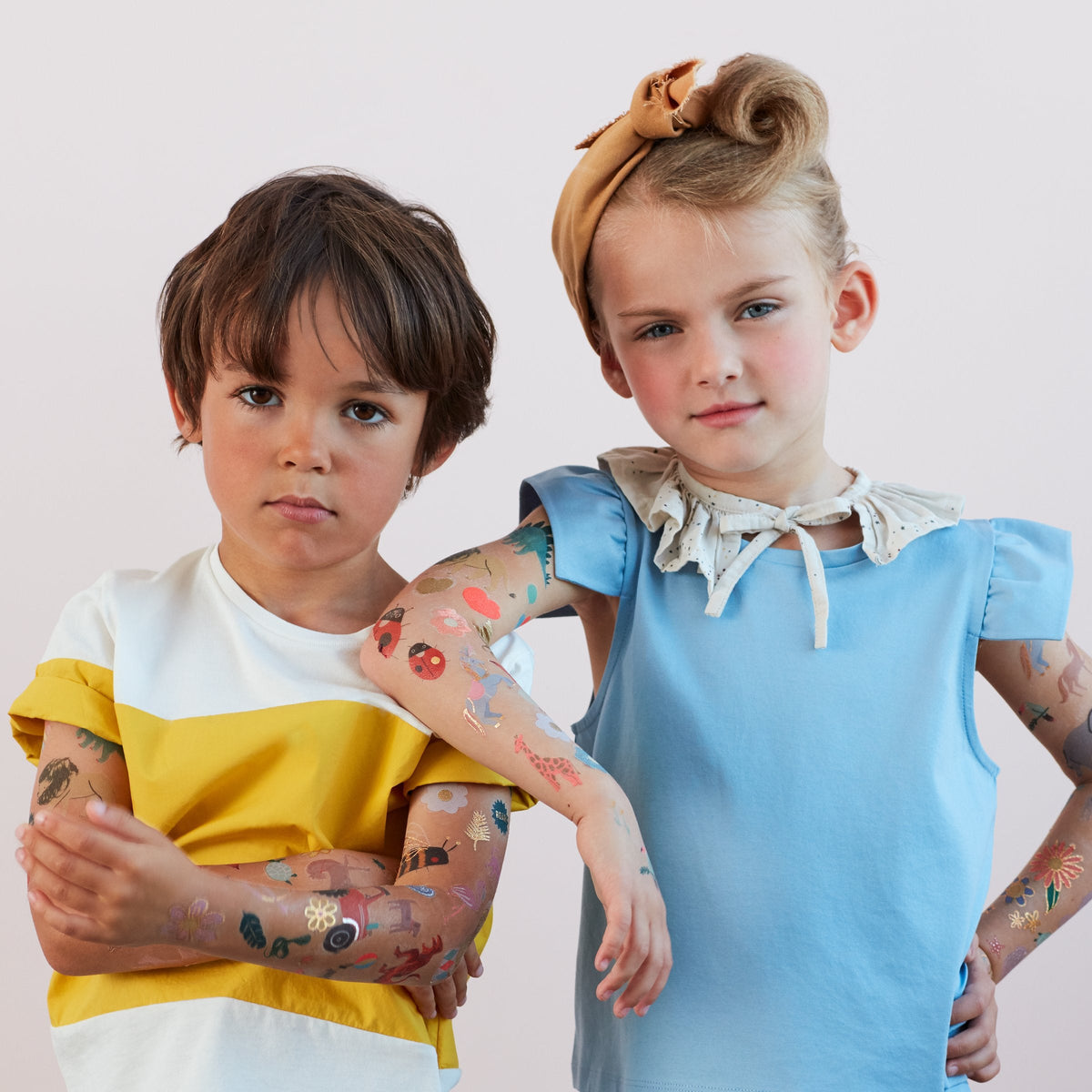 Two children with Meri Meri Large Tattoos, one wearing a yellow and white shirt, the other in a blue top with a headband, standing with arms crossed against a light background. These fun designs by Meri Meri make for an exciting party activity and are a perfect gift for guests.