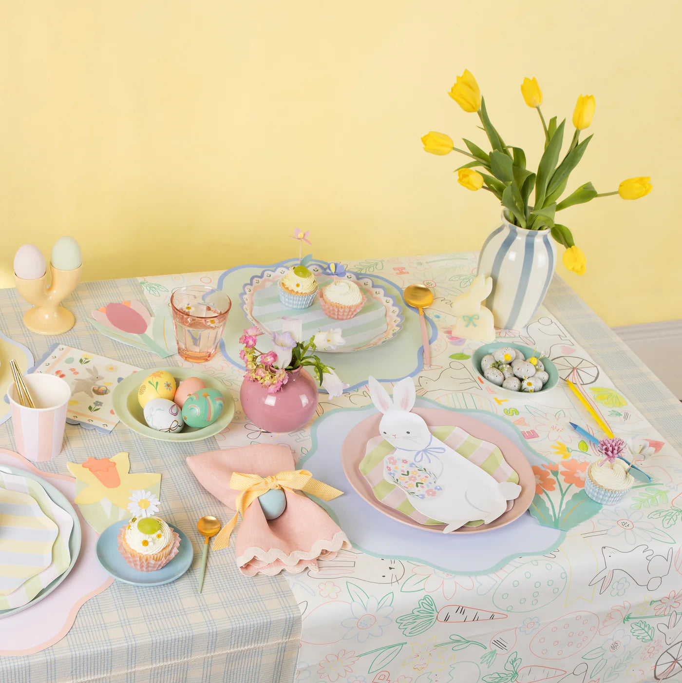 An Easter table setting bursts with ideas, featuring a bunny plate, eggs, cupcakes, and tulips on a Meri Meri Easter Colour In Activity Tablecloth that creatively adds color to your festive feast.