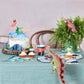 A table set for a dinosaur-themed party features a cake and cupcakes decorated with Meri Meri Cake Toppers from the Meri Meri brand, along with other decorations adorned with various dinosaur designs, creating an exciting prehistoric atmosphere.