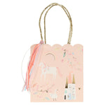 A light pink Meri Meri gift bag with a scalloped top edge, featuring fun illustrations of a unicorn, rainbow, flowers, a castle, and a swan, with gold woven handles and ribbon decorations. Perfect as party bags for any celebration.