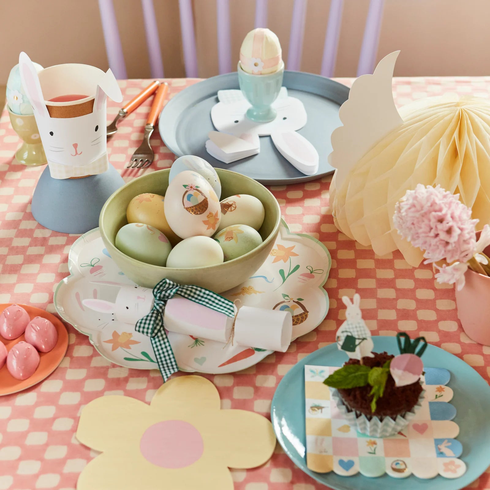 An Easter-themed table showcases painted eggs in a bowl, Meri Meri Lop Eared Bunny Cups, a cupcake, a paper flower, and a checkered tablecloth.