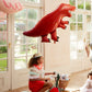 A child wearing a tiara rides a bicycle indoors, eyes gleaming with delight as they gaze at the shiny Meri Meri Foil Balloon shaped like a red dinosaur.