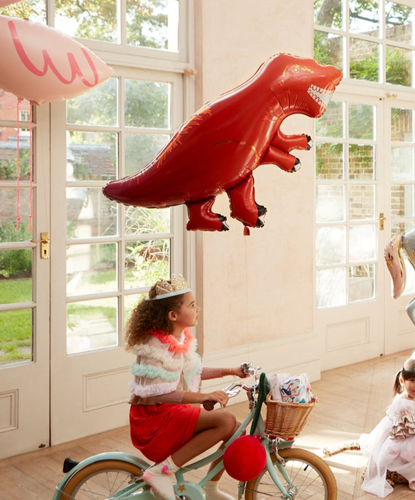 A child wearing a tiara rides a bicycle indoors, eyes gleaming with delight as they gaze at the shiny Meri Meri Foil Balloon shaped like a red dinosaur.