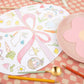 Meri Meri's charming Easter Placemats are egg-shaped and feature pastel drawings of bunnies, carrots, and spring icons. Perfect for Easter coloring enthusiasts, these placemats pair beautifully with a pink flower plate and colored pencils on a checkered tablecloth.