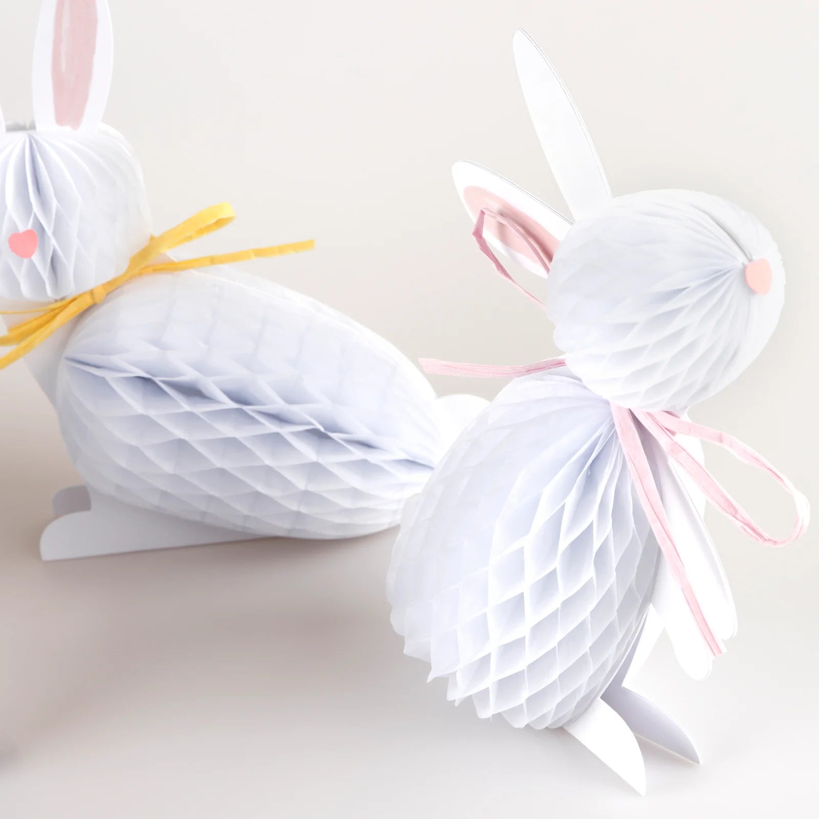 Two Bunny Honeycomb Decorations by Meri Meri, featuring pink and yellow ribbons, hop into a cheerful nursery setting. These charming 3D paper bunnies add a playful touch against the plain background.