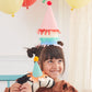 A child wearing a vibrant stack of Meri Meri Party Hats grins while hugging a stuffed dinosaur adorned with a matching hat, surrounded by balloons.