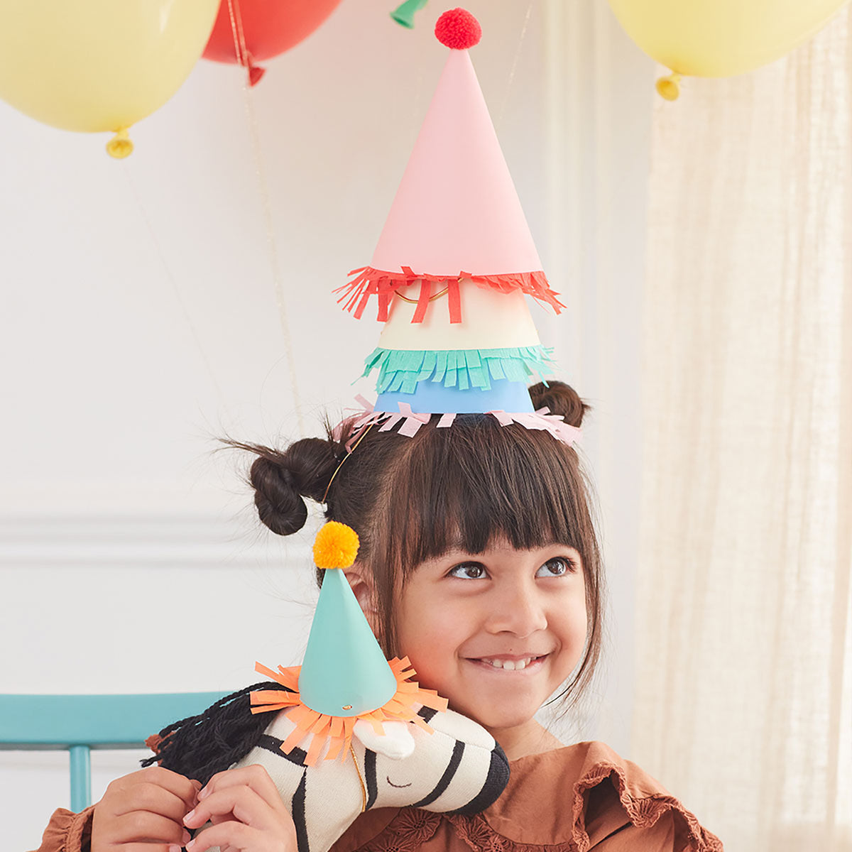 A child wearing a vibrant stack of Meri Meri Party Hats grins while hugging a stuffed dinosaur adorned with a matching hat, surrounded by balloons.