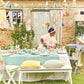 A child arranges an outdoor Easter table with pastel pillows, honeycomb eggs, and flowers. The scene is enhanced by a Meri Meri Easter Garland and a "Happy Easter" banner as a stone wall and lush greenery create the perfect backdrop.