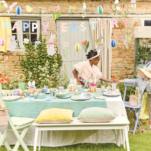 A child arranges an outdoor Easter table with pastel pillows, honeycomb eggs, and flowers. The scene is enhanced by a Meri Meri Easter Garland and a "Happy Easter" banner as a stone wall and lush greenery create the perfect backdrop.