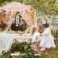 Two children dressed in costumes stand in a garden near an enchanted canopy decorated with the Meri Meri Princess Party Garland by Meri Meri, creating a magical atmosphere filled with flowers and paper crafts reminiscent of a fairy tale.