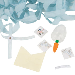 Meri Meri's Surprise Eggs offer assorted craft items ideal for Easter baskets, including light blue streamers, floral and rabbit stickers, a carrot eraser, a plastic egg, a small envelope, and a fortune cookie message.