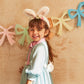 A cheerful child in a blue dress and Meri Meri's Plush Bunny Ears grins before a pastel bow garland, holding a gingham bag.