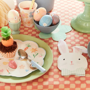 An Easter table adorned with pastel eggs, floral cups, a carrot cupcake, bunny decor, and a decorative spoon comes alive on a pink checkered cloth accented by Meri Meri Easter Bunny Napkins.