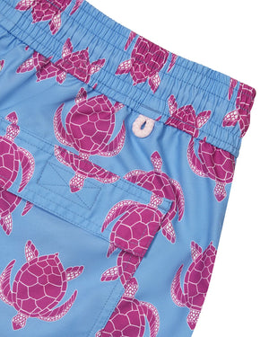 Close-up of the Turtle Trunks from Pink House Mustique, showcasing blue swim shorts with a playful pink sea turtle pattern and an elastic waistband. Designed with a buttoned pocket and crafted from quick-dry recycled fabric, these stylish swim trunks combine comfort and sustainability for your days by the water.