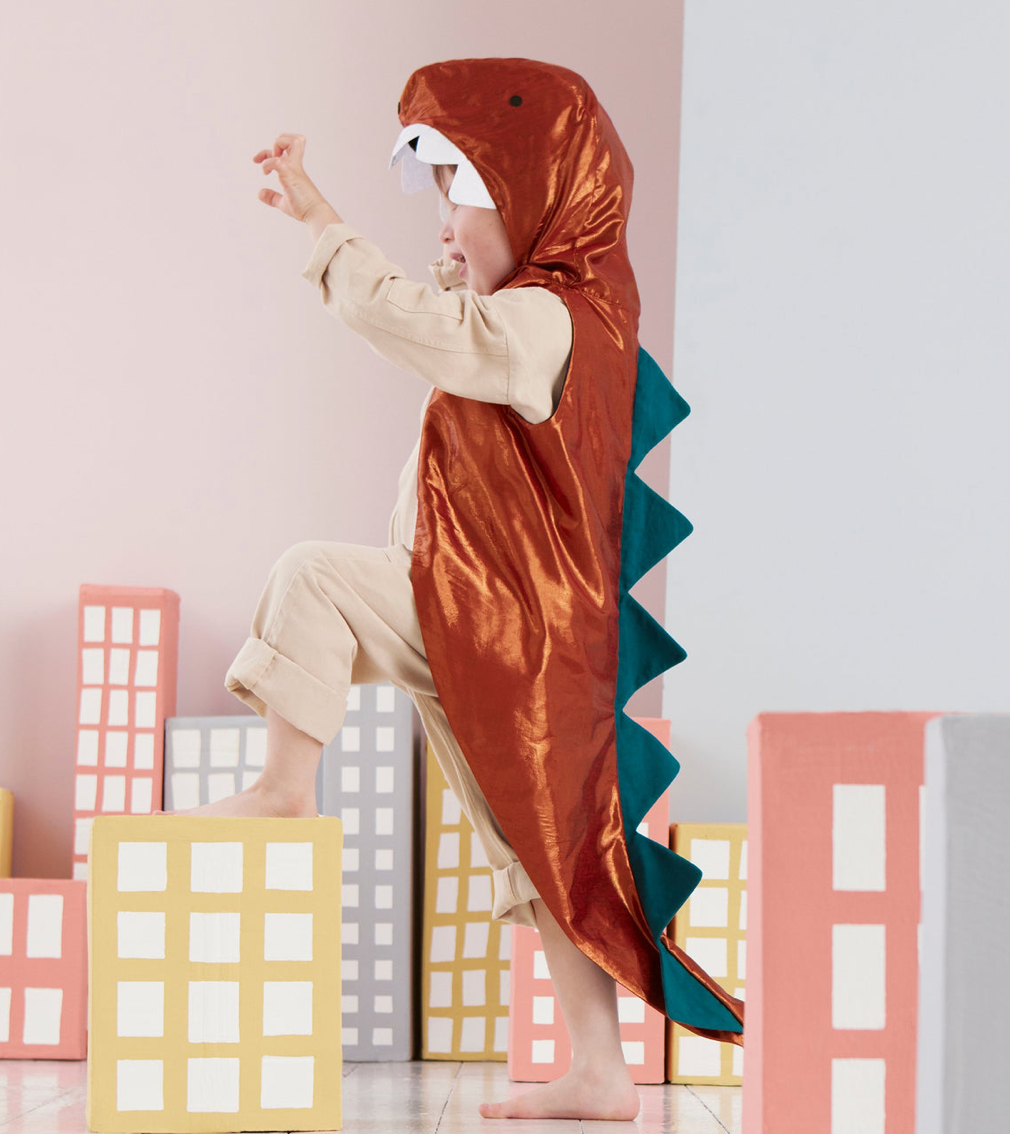 A child dressed in a Meri Meri Dinosaur Costume, complete with a hooded jacket adorned with spikes, is standing on one leg and posing playfully amid cardboard cutout buildings.