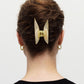 A person with curly brown hair is seen from the back, wearing a black top. Their hair is secured with a large, light-colored Machete Midi Heirloom Claw made from Italian acetate, and they have small gold hoop earrings.