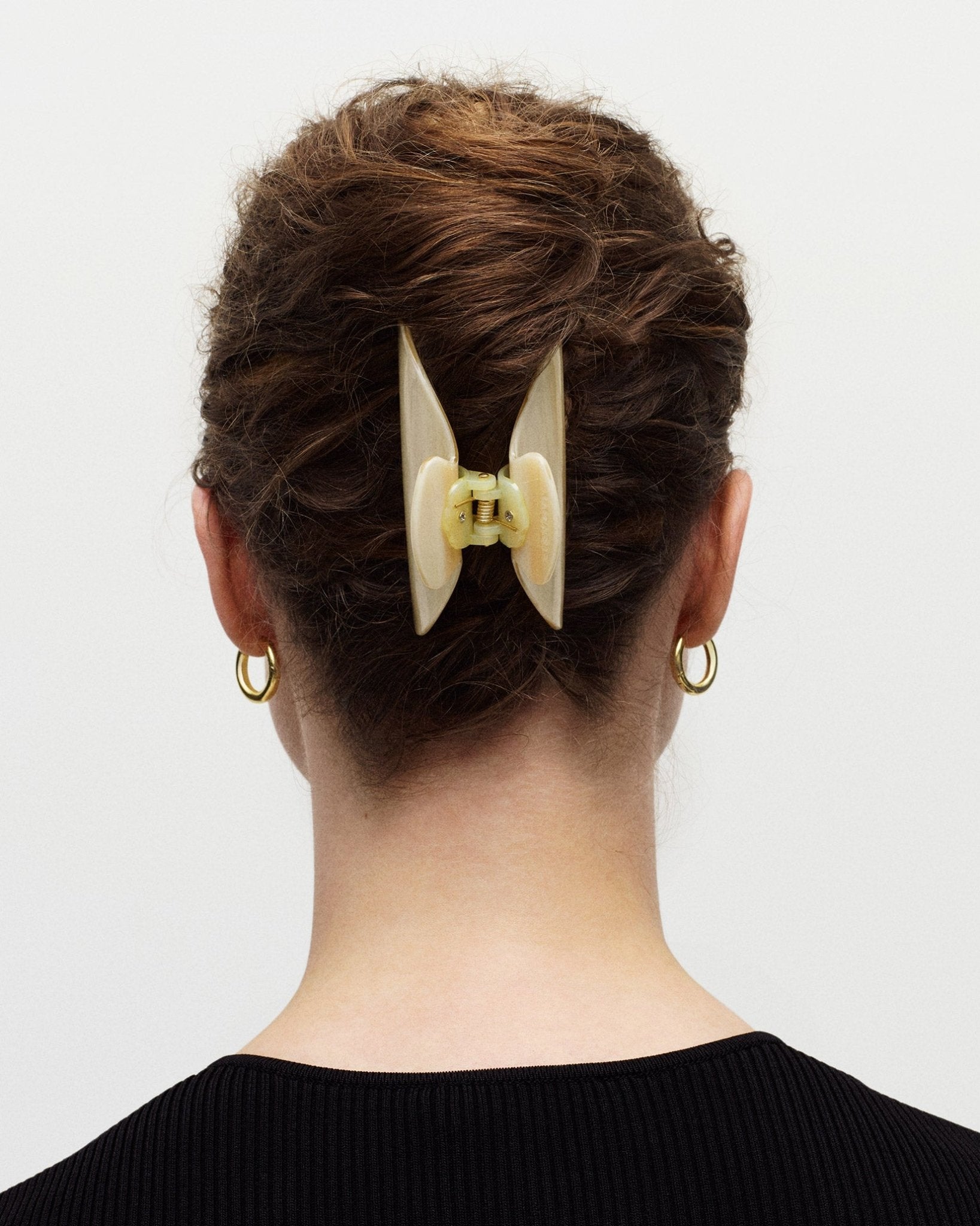 A person with curly brown hair is seen from the back, wearing a black top. Their hair is secured with a large, light-colored Machete Midi Heirloom Claw made from Italian acetate, and they have small gold hoop earrings.