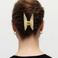 A person with brown hair styled in a twist secured by the Machete Midi Heirloom Claw in cream Italian acetate, wearing gold hoop earrings and a black top, is photographed from behind against a plain background.