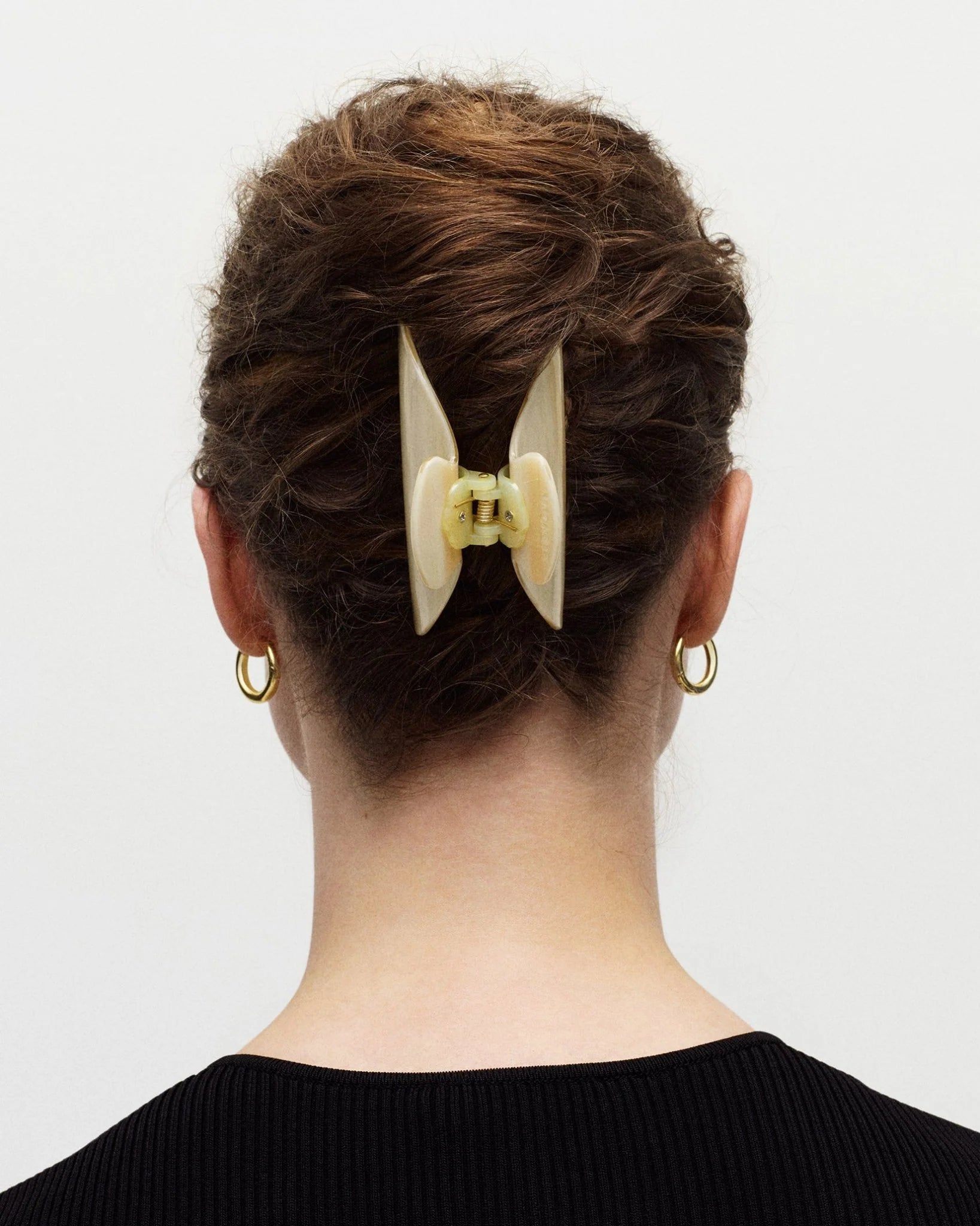 A person with brown hair styled in a twist secured by the Machete Midi Heirloom Claw in cream Italian acetate, wearing gold hoop earrings and a black top, is photographed from behind against a plain background.