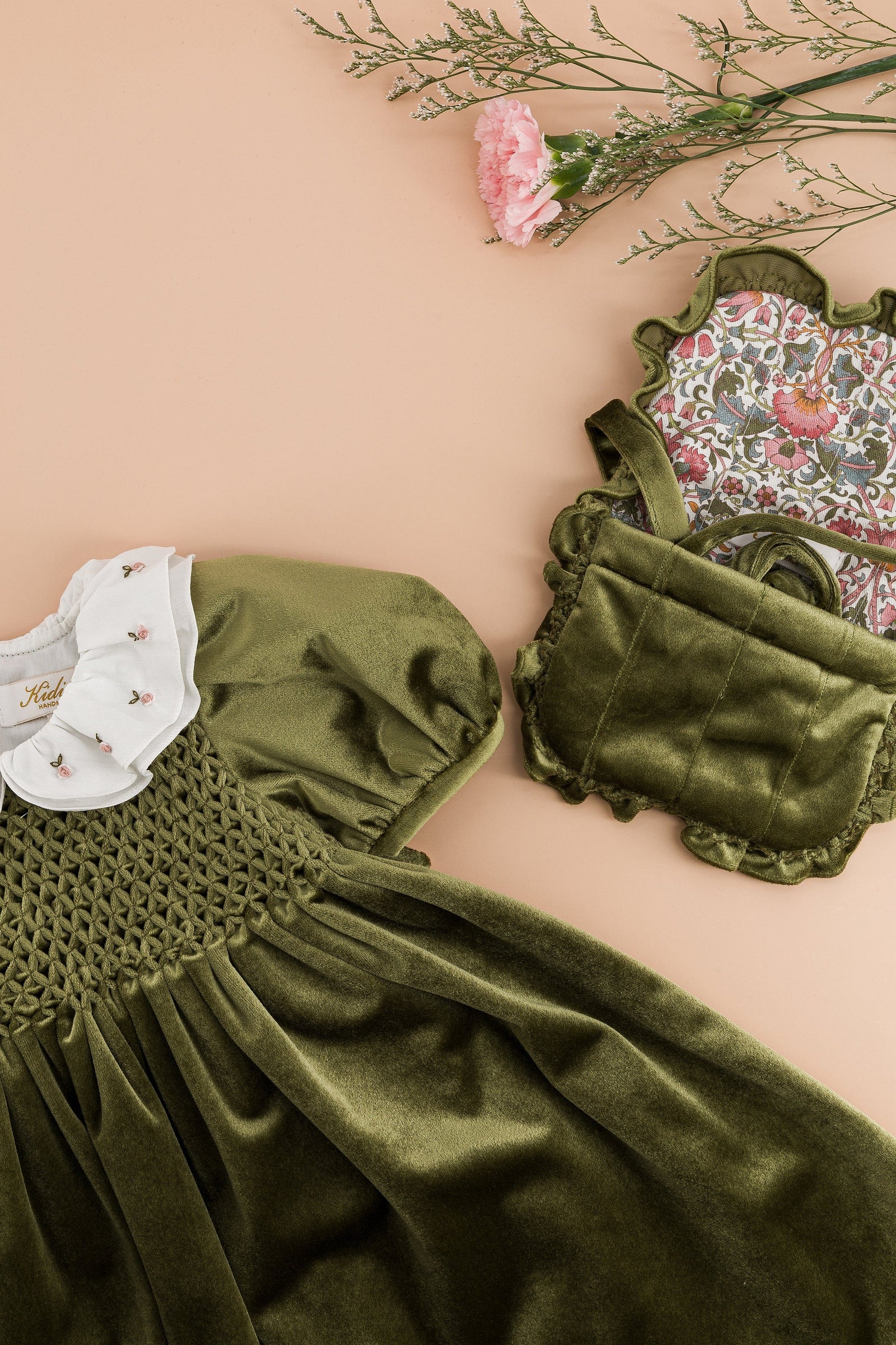 Green olive velvet dress with puffed sleeves, white embroidered collar, Kidiwi’s Mimosa Smocked Shoulder Bag, floral patterned item, and pink carnation on beige—featured in the winter 2025 collection.