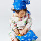 A baby dressed in an Arsene & Les Pipelettes organic cotton Baby Sweatshirt, wearing a blue bandana and patterned pajamas, sits on the floor looking down and touching a blue quilted blanket with a yellow patch.
