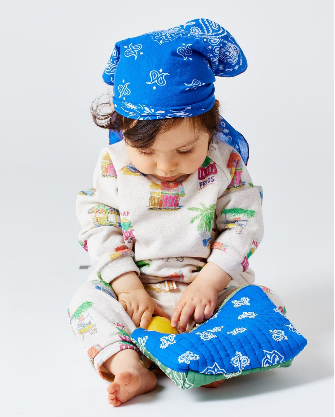 A baby dressed in an Arsene & Les Pipelettes organic cotton Baby Sweatshirt, wearing a blue bandana and patterned pajamas, sits on the floor looking down and touching a blue quilted blanket with a yellow patch.