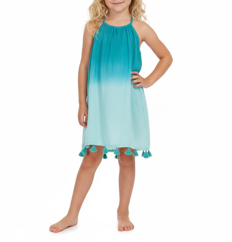 A young child models the Mer Girls’ Mini Chantal Dress, a sleeveless blue ombre summer dress featuring tassels at the hem, while standing barefoot against a plain white background.