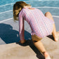 A young girl wearing the Minnow Girls' Floral Tile Rashguard One Piece kneels at the pool's edge, leaning forward with her hands near the water.