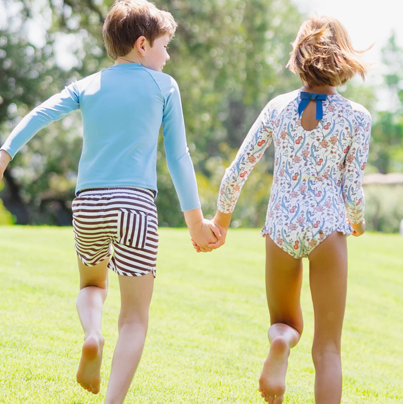 Two girls in Minnow Girls' Feathered Floral Rashguard One Piece hold hands and run barefoot on green grass, with blurred trees in the background.