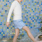 A child wearing a long-sleeve white shirt and Minnow Boys' Starburst Boardie with an elastic waistband stands in shallow water before a colorful tiled wall, splashing playfully with one foot.