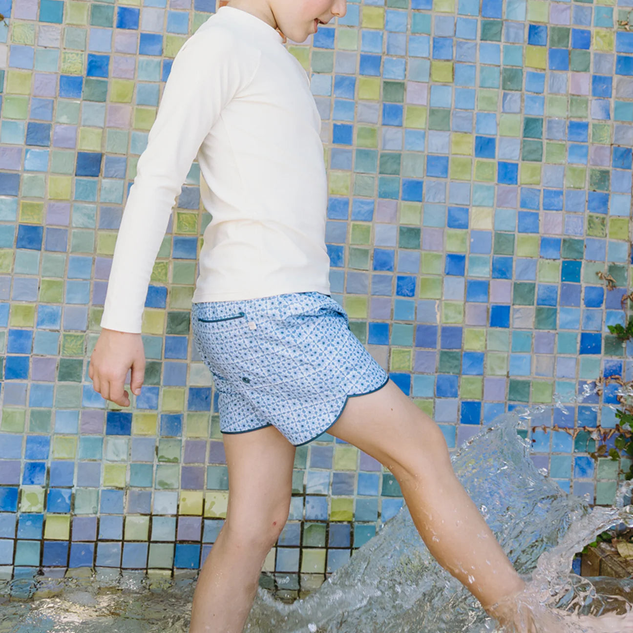 A child wearing a long-sleeve white shirt and Minnow Boys' Starburst Boardie with an elastic waistband stands in shallow water before a colorful tiled wall, splashing playfully with one foot.