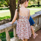 A young girl with a long braid stands on a stone balcony, wearing the Minnow Girls' Feathered Floral Square Neck Strap Dress, looking out at a green garden.