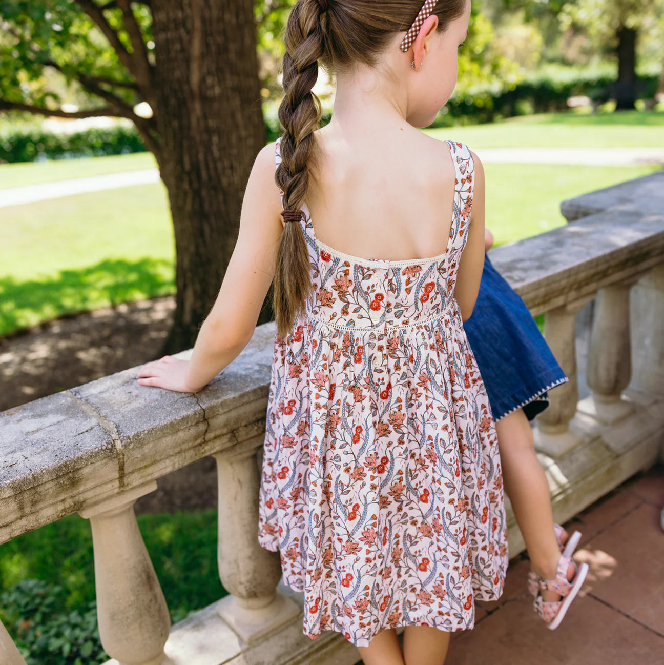 A young girl with a long braid stands on a stone balcony, wearing the Minnow Girls' Feathered Floral Square Neck Strap Dress, looking out at a green garden.