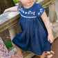 A young girl in the Minnow Girl's Chambray Embroidered Cap Sleeve Dress sits on a stone railing outdoors.